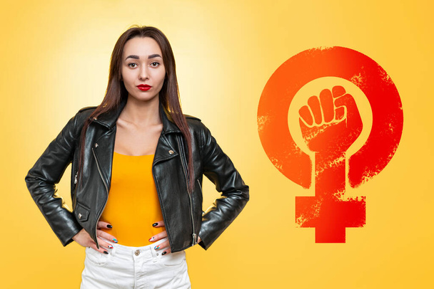 Концепція рівності та фемінізму. Кавказька жінка в шкіряній куртці впевнено стоїть на жовтому тлі. Знак жіночої сили.. - Фото, зображення