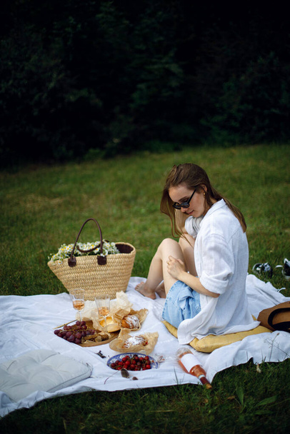 a young woman sits on a white mat in a park or garden and holds a glass of rose wine in her hand. girl waiting for a friend for a picnic in the garden. picnic with cheese, fruits and wine - Photo, Image