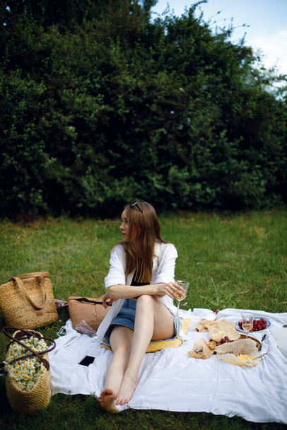 молодая женщина сидит на белом коврике в парке или саду и держит в руке бокал розового вина. Девушка ждет друга для пикника в саду. пикник с сыром, фруктами и вином - Фото, изображение