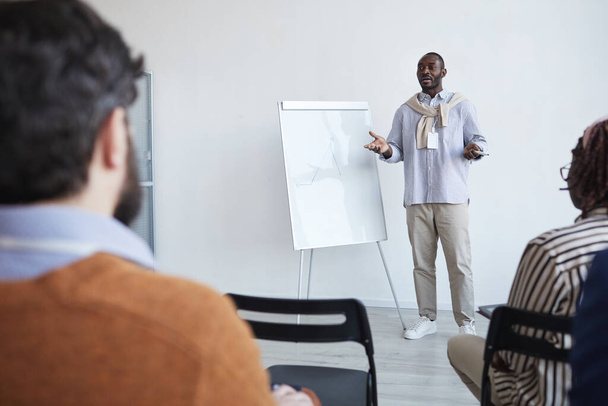 Weitwinkelporträt eines afroamerikanischen Business-Coaches, der auf Konferenzen oder Bildungsseminaren mit dem Publikum spricht, während er am Whiteboard steht und gestikuliert, Kopierraum - Foto, Bild