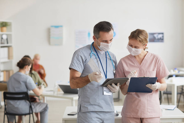 Portrait taille haute de deux médecins portant des masques et parlant tout en regardant la tablette dans la clinique médicale, copiez l'espace - Photo, image