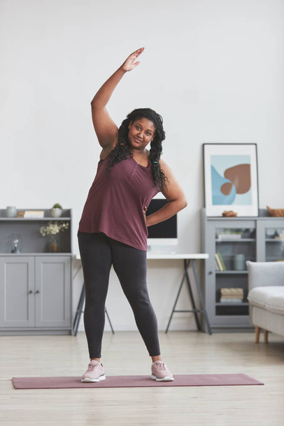 Retrato de comprimento total vertical de mulher afro-americana curvilínea trabalhando em casa e sorrindo para a câmera enquanto esticando os braços em pé no tapete de ioga no interior mínimo, espaço de cópia - Foto, Imagem