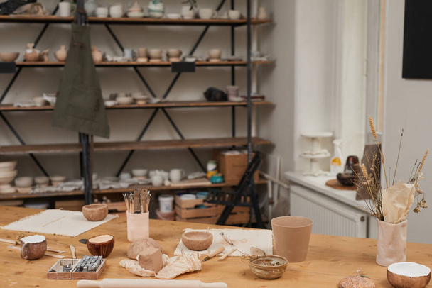 Imagen de fondo en tonos cálidos del taller de cerámica vacía con mesa de madera y herramientas en primer plano, espacio para copiar - Foto, Imagen