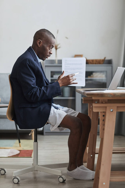 Vertical vista lateral retrato de homem afro-americano vestindo jaqueta formal e shorts durante reunião on-line enquanto trabalhava em casa - Foto, Imagem