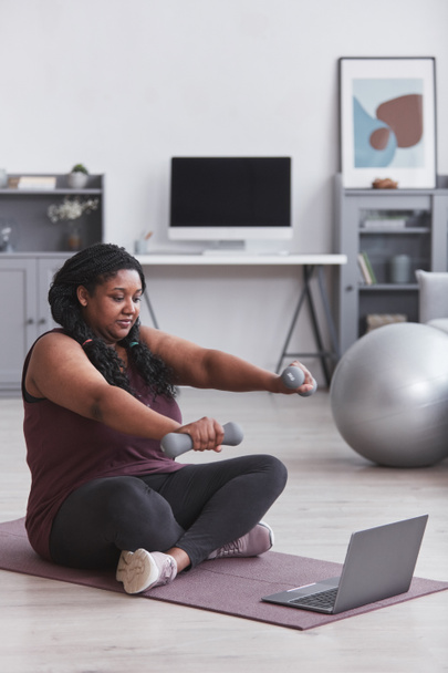Retrato de longitud vertical de una mujer afroamericana con curvas haciendo ejercicio en casa con pesas mientras está sentada en una esterilla de yoga y viendo videos de entrenamiento en línea, espacio para copiar - Foto, Imagen