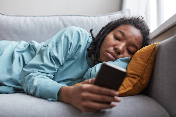Portrét depresivní Afroameričanky držící smartphone při ležení na pohovce doma se zaměřením na nedokonalosti pleti - Fotografie, Obrázek
