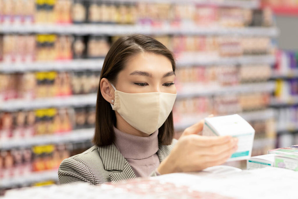 Young contemporary elegant female consumer in protective mask holding box with face cream while choosing one for herself in supermarket - Foto, Bild