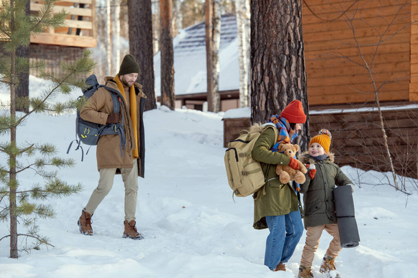 Młody człowiek z plecakiem podążający za żoną i córeczką w kierunku wiejskiego domu po przyjeździe do nowoczesnego ośrodka zimowego - Zdjęcie, obraz