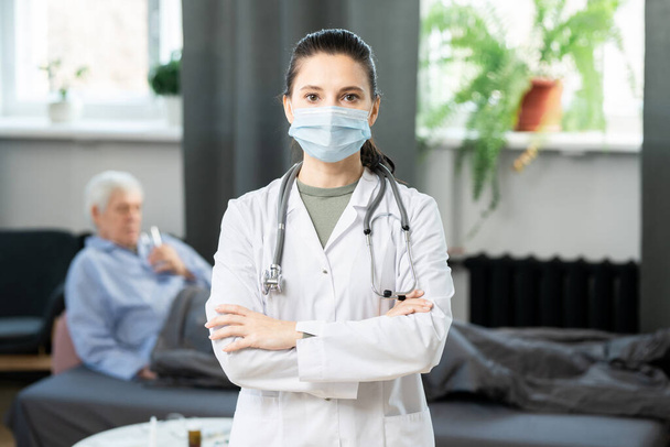 Young general practitioner in whitecoat and protective mask crossing her arms by chest and looking at you against sick senior patient in bed - Photo, Image