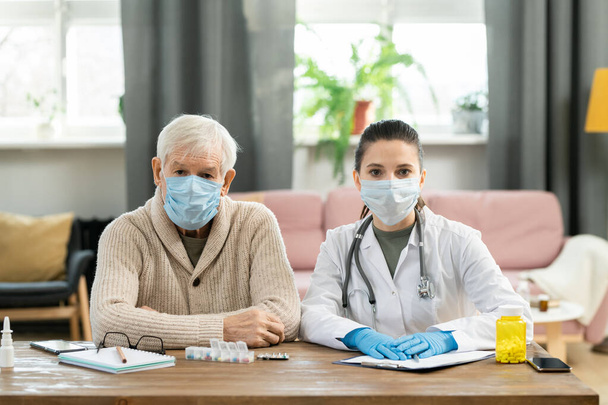 Nemocný starší muž pacient a mladá brunetka lékař v ochranných maskách sedí vedle stolu vedle sebe a dívá se na vás - Fotografie, Obrázek