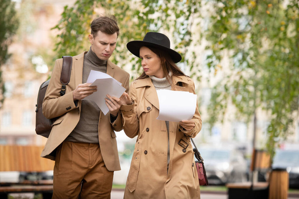 Dwóch młodych poważnych ekonomistów w inteligentnych ubraniach codziennych przeglądających gazety i dyskutujących o nich w parku podczas przerwy lub po pracy - Zdjęcie, obraz