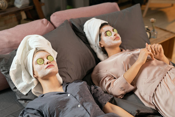 Mujeres jóvenes en pijama de seda, toallas en la cabeza, máscaras de barro en la cara y rodajas de pepino en los ojos cerrados disfrutando de un procedimiento de belleza en la cama - Foto, Imagen