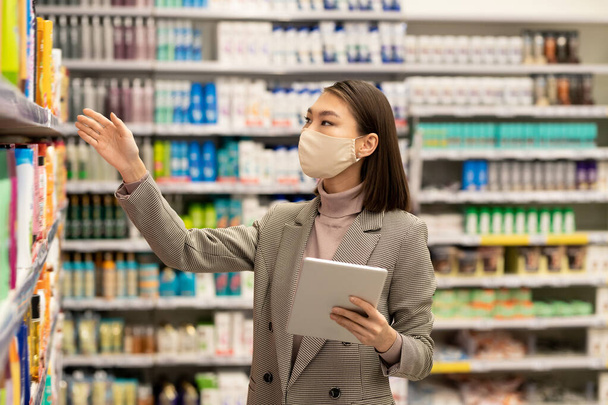 Young Asian female consumer with tablet standing by shelf with bodycare products and choosing one for herself while using touchpad - Photo, Image