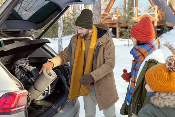 Kortárs fiatal, három fős család meleg télikabátban, akik kiveszik a csomagjaikat a csomagtartóból, miközben az autó hátuljában állnak utazás közben. - Fotó, kép