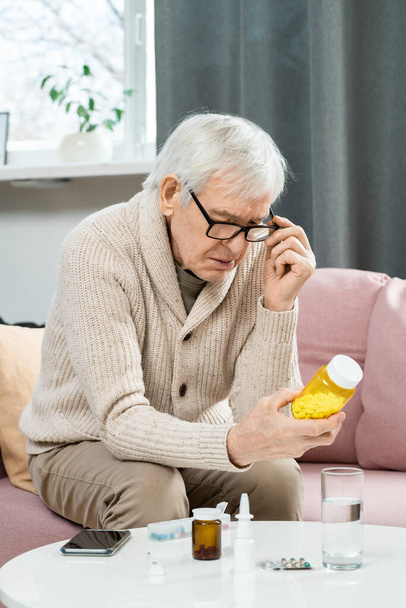 Älterer Mann in Freizeitkleidung und Brille sitzt auf Sofa im Wohnzimmer, hält Tablettenflasche über Tisch und liest Tabletten - Foto, Bild