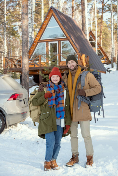 Young cheerful couple with backpacks standing in front of camera against their car and country house and looking at you on winter weekend - Photo, image