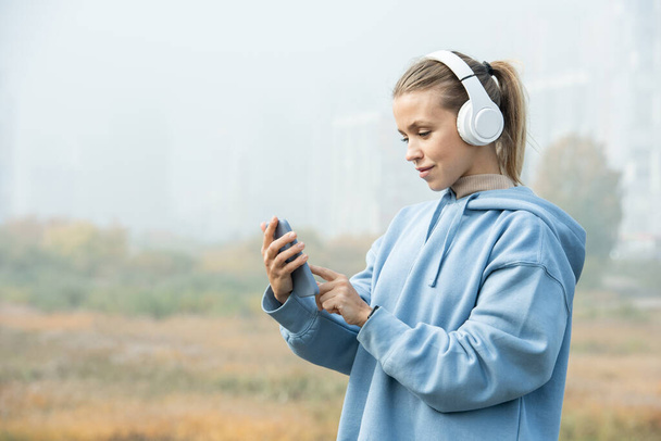 Młoda blondynka w białych słuchawkach i niebieskim kapturze słuchająca muzyki i wskazująca na ekran smartfona przed kamerą - Zdjęcie, obraz