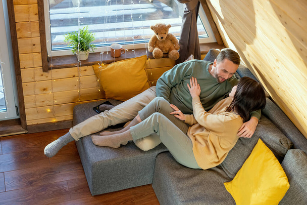 Felices jóvenes relajados marido y mujer en ropa de casualyacente en un gran sofá dentro de su casa de campo, disfrutando de fin de semana y charlando - Foto, Imagen