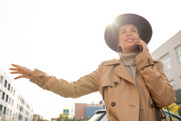 Молодая элегантная улыбающаяся деловая женщина в бежевом пальто и черной шляпе пытается поймать такси, стоя на дороге в городской среде - Фото, изображение
