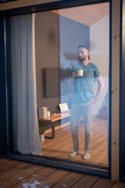 Молодий бородатий чоловік у повсякденному одязі тримає кухоль з гарячим напоєм і дивиться через велике вікно, стоячи на дерев'яній підлозі у вітальні
 - Фото, зображення