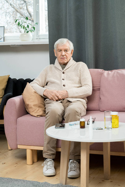 Hombre mayor enfermo en ropa casual sentado en el sofá por una pequeña mesa con medicamentos y un vaso de agua y esperando al médico en la sala de estar - Foto, imagen
