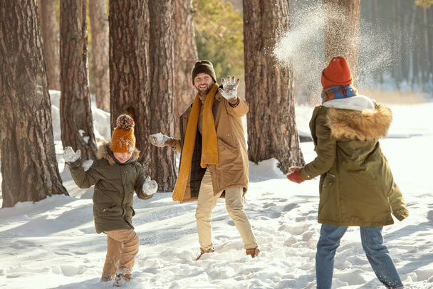 Χαρούμενος νεαρός άνδρας, η γυναίκα του και η χαριτωμένη κορούλα τους με ζεστά χειμωνιάτικα ρούχα να παίζουν χιονόμπαλες ενώ διασκεδάζουν στο χιόνι - Φωτογραφία, εικόνα
