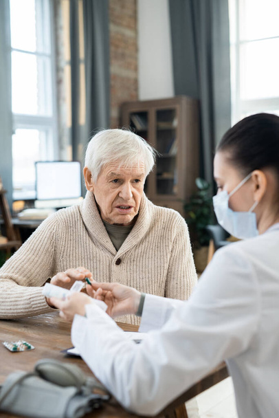 Fiatal női orvos fehérköpenyben és védőmaszkban tablettákkal teli tartályt ad át egy beteg idős férfinak, aki leírja neki a tüneteit. - Fotó, kép