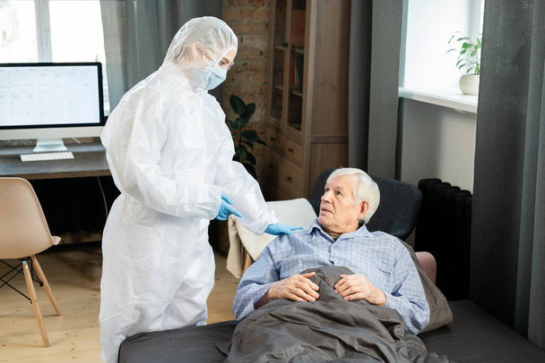 Junge Ärztin in Schutzanzügen berührt kranken Senior auf Couch unter Decke und gibt ihm ärztlichen Rat - Foto, Bild