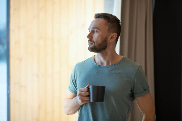Junger bärtiger, entspannter Mann im grauen T-Shirt hält einen Becher mit heißem Tee oder Kaffee in der Hand und schaut durch ein großes Fenster, während er zu Hause bleibt - Foto, Bild