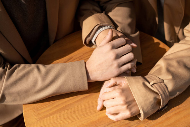 Mãos de homem e mulher afetuosos jovens em casacos de traquete bege sentados à mesa de madeira redonda pequena perto um do outro no dia ensolarado - Foto, Imagem