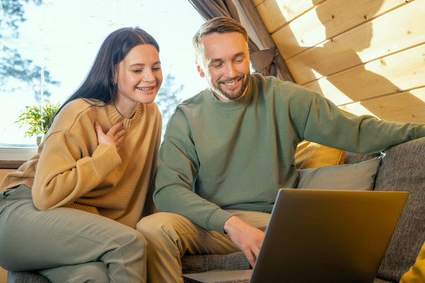Feliz joven pareja cariñosa en ropa de casualwear sentado en un gran sofá delante de la pantalla del ordenador portátil y la comunicación a través de chat de vídeo - Foto, imagen