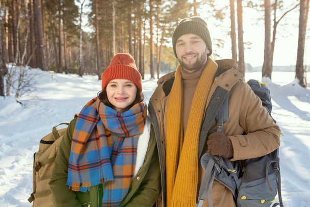 Joven pareja sonriente en ropa de invierno caliente de pie delante de la cámara contra la nieve y el bosque mixto y mirándote en un día soleado - Foto, Imagen