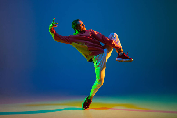 Bailarín masculino joven bailando hip-hop aislado sobre fondo azul en luz de neón - Foto, Imagen