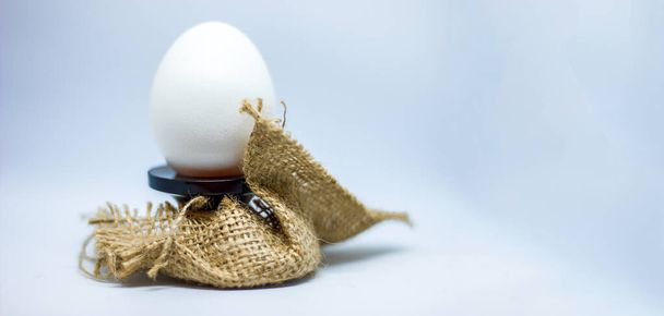 białe jajko na podstawce ozdobionej płótnem w formie gniazda na niebieskim tle, temat Wielkanocy i zdrowego odżywiania białek, przestrzeń do kopiowania tekstu - Zdjęcie, obraz