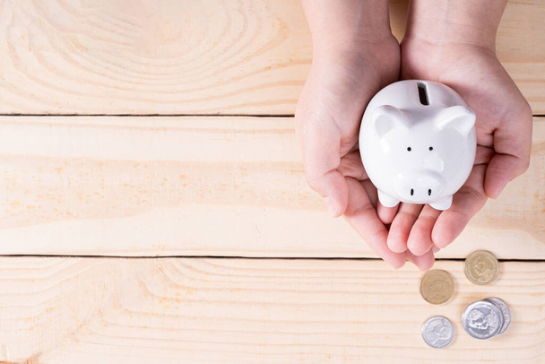 Hands hold a piggy bank with a pile of coins on wooden background. Saving money and investment concept. - Zdjęcie, obraz