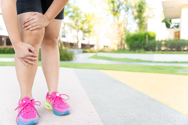 Жирная женщина бегунья с травмой ноги и болью. Руки хватаются за больное колено во время пробежки по парку. - Фото, изображение