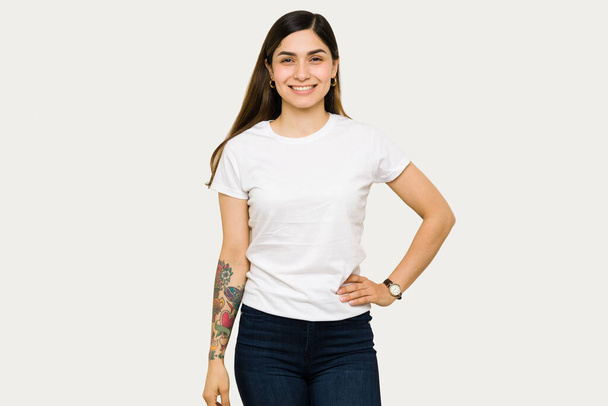 Ευτυχισμένη νεαρή γυναίκα που ποζάρει με το ένα χέρι στο γοφό της και χαμογελάει. Γυναίκα φορώντας ένα mock up t-shirt σχεδιασμό, ενώ στέκεται μπροστά από ένα λευκό φόντο - Φωτογραφία, εικόνα