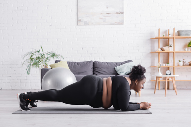 Seitenansicht einer afrikanisch-amerikanischen Plus-Size-Frau in Sportbekleidung beim Planken im Wohnzimmer - Foto, Bild