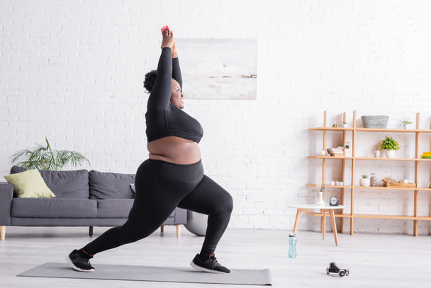 повна довжина весела афроамериканка плюс розмір жінка в спортивному одязі, стоячи в позі йоги у вітальні
 - Фото, зображення
