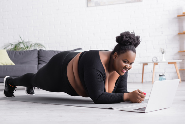 glückliche afrikanisch-amerikanische Plus-Size-Frau beim Online-Sporttraining auf dem Laptop beim Planken im Wohnzimmer - Foto, Bild