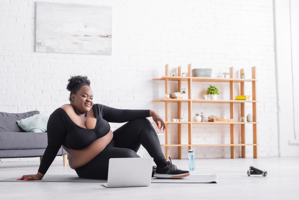 uśmiechnięta Afroamerykanka plus size kobieta oglądająca trening online na laptopie siedząc na macie fitness  - Zdjęcie, obraz