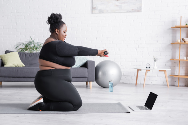 вид сбоку на африканскую женщину plus size, наблюдающую онлайн-тренировку на ноутбуке во время упражнений с гантелями на фитнес-коврике  - Фото, изображение