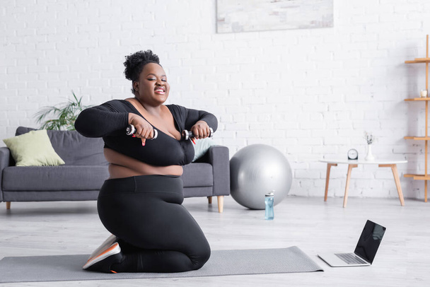 lächelnde afrikanisch-amerikanische Plus-Size-Frau, die in der Nähe von Laptop und Fitnessmatte mit Kurzhanteln trainiert  - Foto, Bild