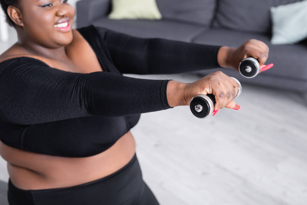 Bulanık Afrikalı Amerikalı, spor giyim bölümünde beden ölçülerinde kadın oturma odasında dambıllarıyla egzersiz yapıyor. - Fotoğraf, Görsel