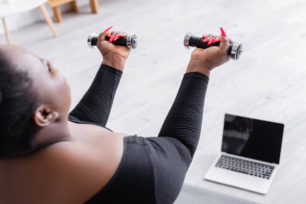 sfocato africano americano plus size donna in abbigliamento sportivo esercizio con manubri vicino computer portatile in soggiorno - Foto, immagini