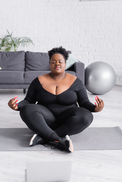 afrikanisch-amerikanische Plus-Size-Frau in Sportbekleidung sitzt in Yoga-Pose neben Laptop im Wohnzimmer - Foto, Bild