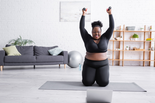 feliz afro-americana oversized mulher em sportswear exercício com halteres perto de laptop  - Foto, Imagem