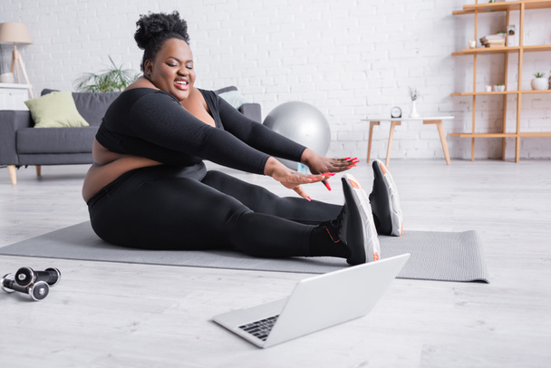 ευτυχισμένη Αφροαμερικανή συν μέγεθος γυναίκα στα αθλητικά που τεντώνεται στο στρώμα φυσικής κατάστασης και ψάχνει στο laptop  - Φωτογραφία, εικόνα