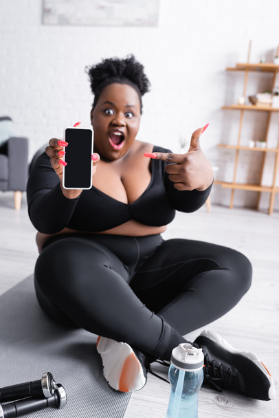 schockierte afrikanisch-amerikanische Plus-Size-Frau in Sportbekleidung zeigt mit dem Finger auf Smartphone mit leerem Bildschirm - Foto, Bild