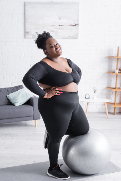 alegre afroamericano más tamaño mujer en ropa deportiva apoyada en la pelota de fitness - Foto, imagen